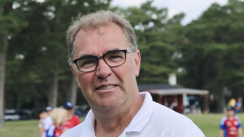 Mikael Nilsson står utomhus framför några träd och tittar in i kameran. Bakom honom pågår Stångaspelen.