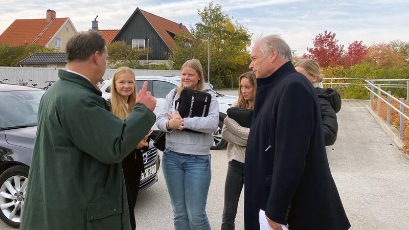Två män står utomhus och samtalar med ett gäng unga elever..