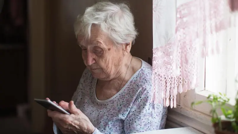 Äldre kvinna sitter med en mobiltelefon i sin hand intill ett fönster.