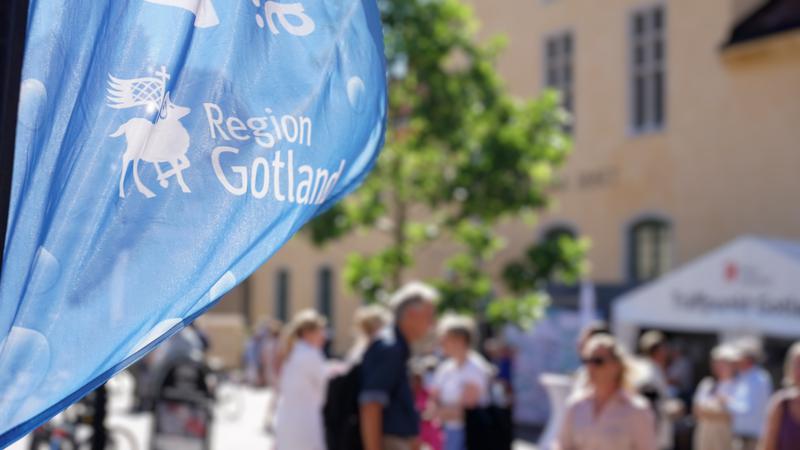 Vajande blå beachflagga med Region Gotlads logotyp och oskarpa personer på ett torg i bakgrunden