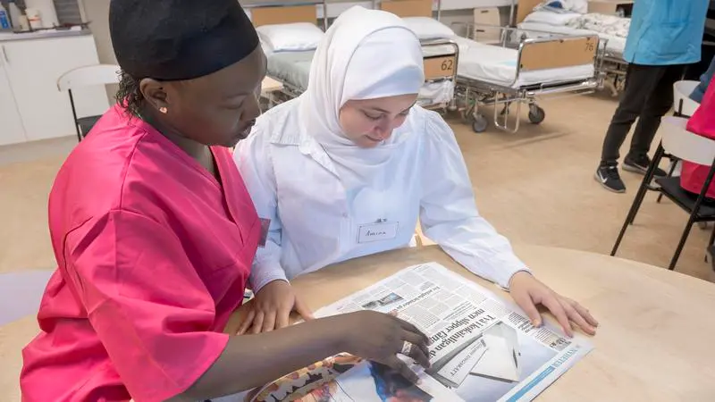 En kvinna i vårdkläder och en kvinna i slöja läser tidningen tillsammans.