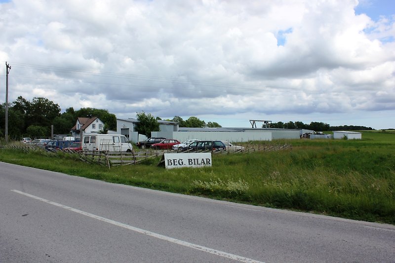 På höger sidan av vägen står det en skylt med begagnade bilar på, därefter ser man tiotals parkerade bilar.