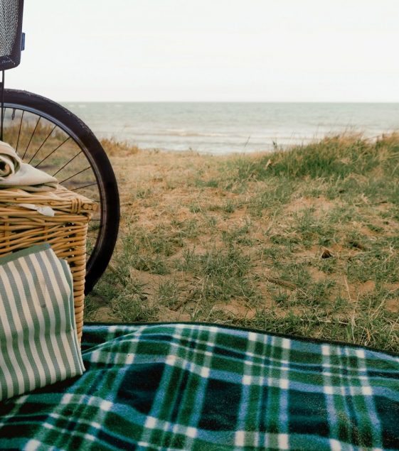 Cykelutflykt på sommarstrand. Picknick filt och korg ligger utplacerad på sandstrand vid havet. En cykel står parkerad bredvid.