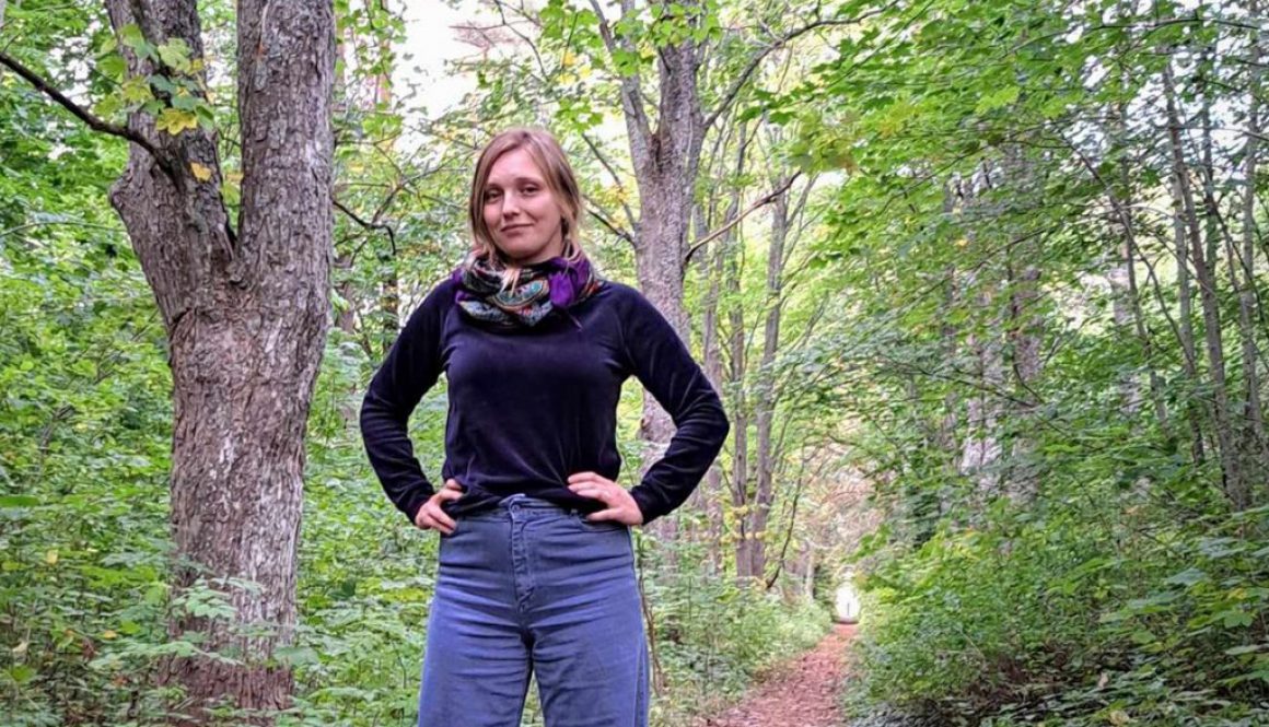 Kvinna står med armarna vilande på höften, Grönskande skog och en lövad stig i bakgrunden