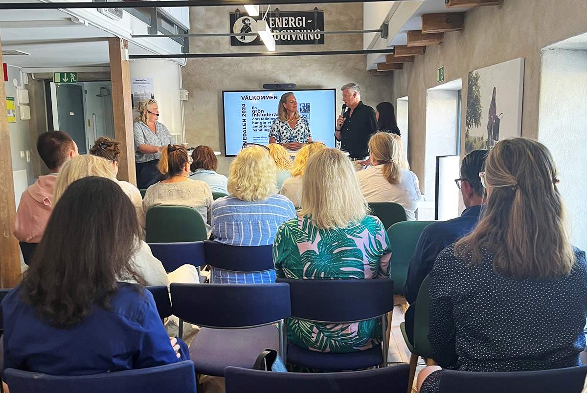 Presentation med tre personer framme på scen fram för en sittande publik