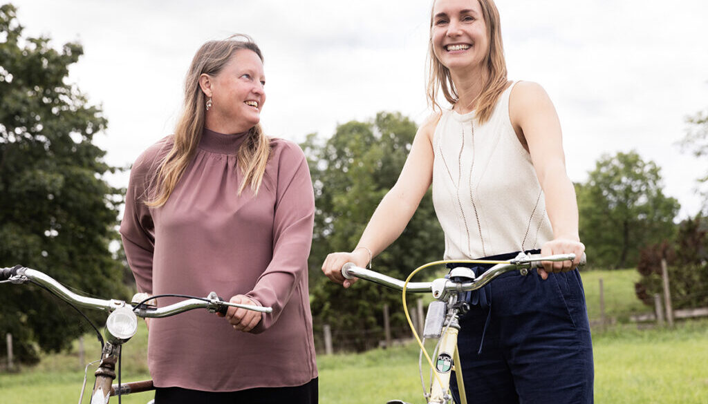 Två leende kvinnor leder sina cyklar omgivna av grönska