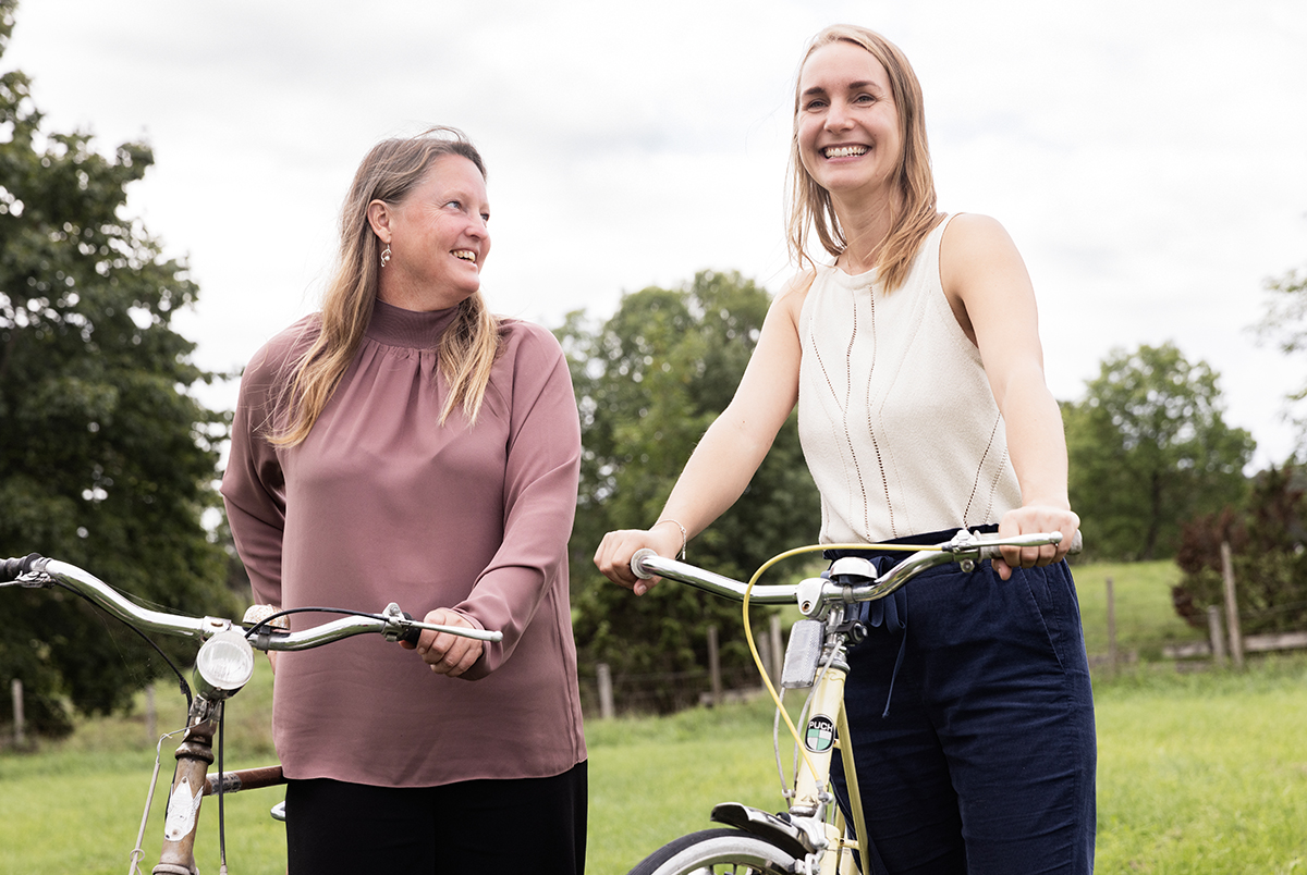 Två leende kvinnor leder sina cyklar omgivna av grönska