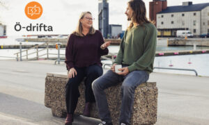 två personer sitter på en bänk i Visby hamn och pratar. Logo Ö-drifta uppe i vänster hörn.