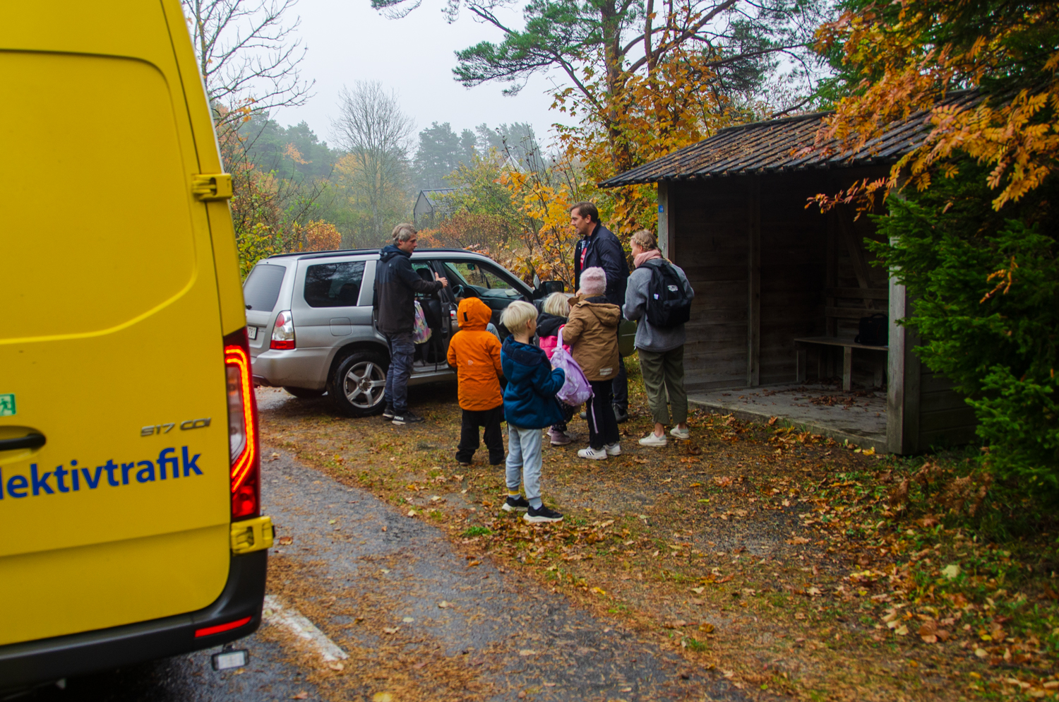 Barn och vuxna har klivit av bussen och möts vid en busskur. Höstväder